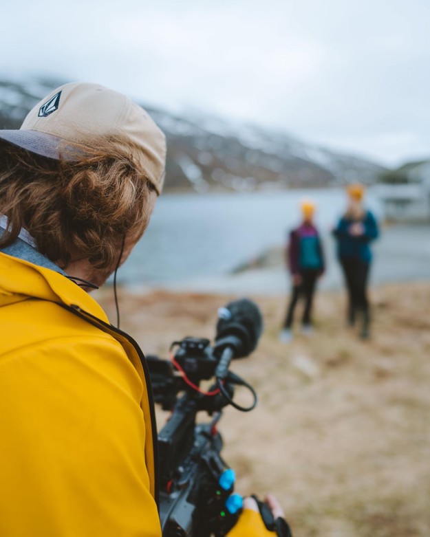 En fotograf i kledd gul jakke og caps bakfrem filmer to jenter langt vekk. de er uklare. 